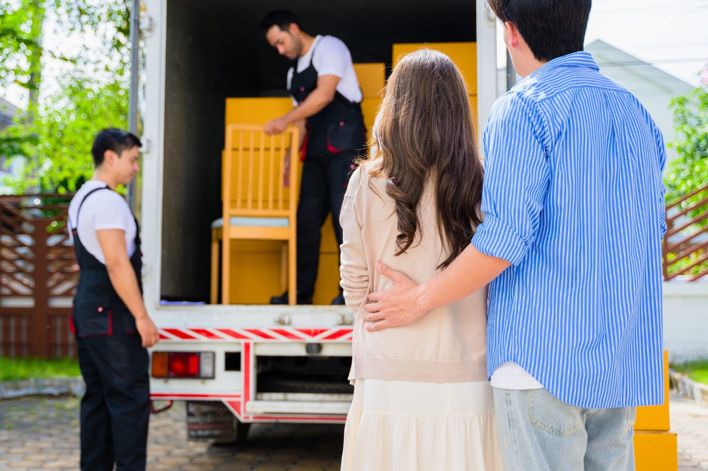 Couple watching professional movers load furniture into a moving truck outside their home.