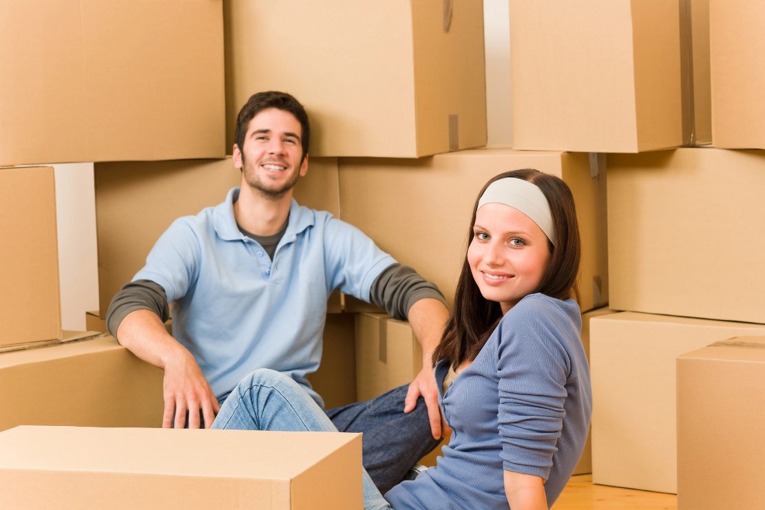 Couple sitting together surrounded by packed cardboard moving boxes.